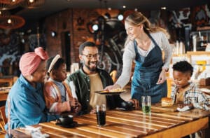 Family, lunch and service of waiter at restaurant with plate, fun and eating at diner together. Customer, table and server at cafe with food for happy African parents, children and relax at gathering